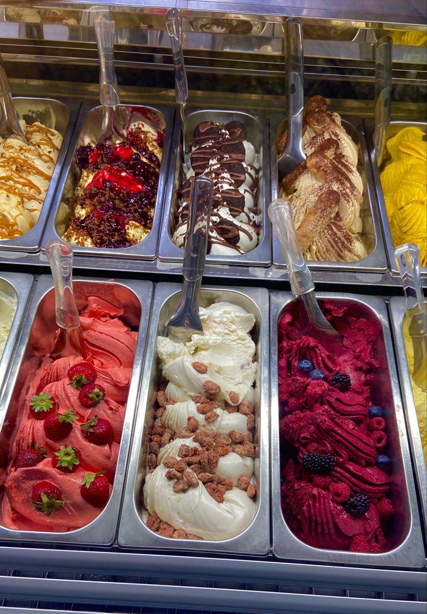 Colorful gelato display with a variety of flavors including strawberry, vanilla, chocolate, and berry, topped with fresh fruit, caramel, and nuts in a metal freezer case.